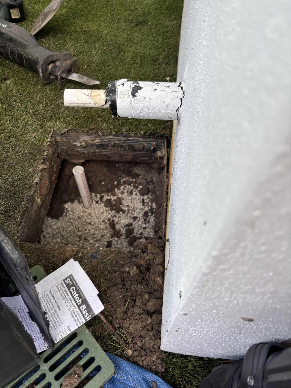Small hand-dug pit in a lawn with exposed white conduit, stone bedding and a drain grate beside it in Delray Beach