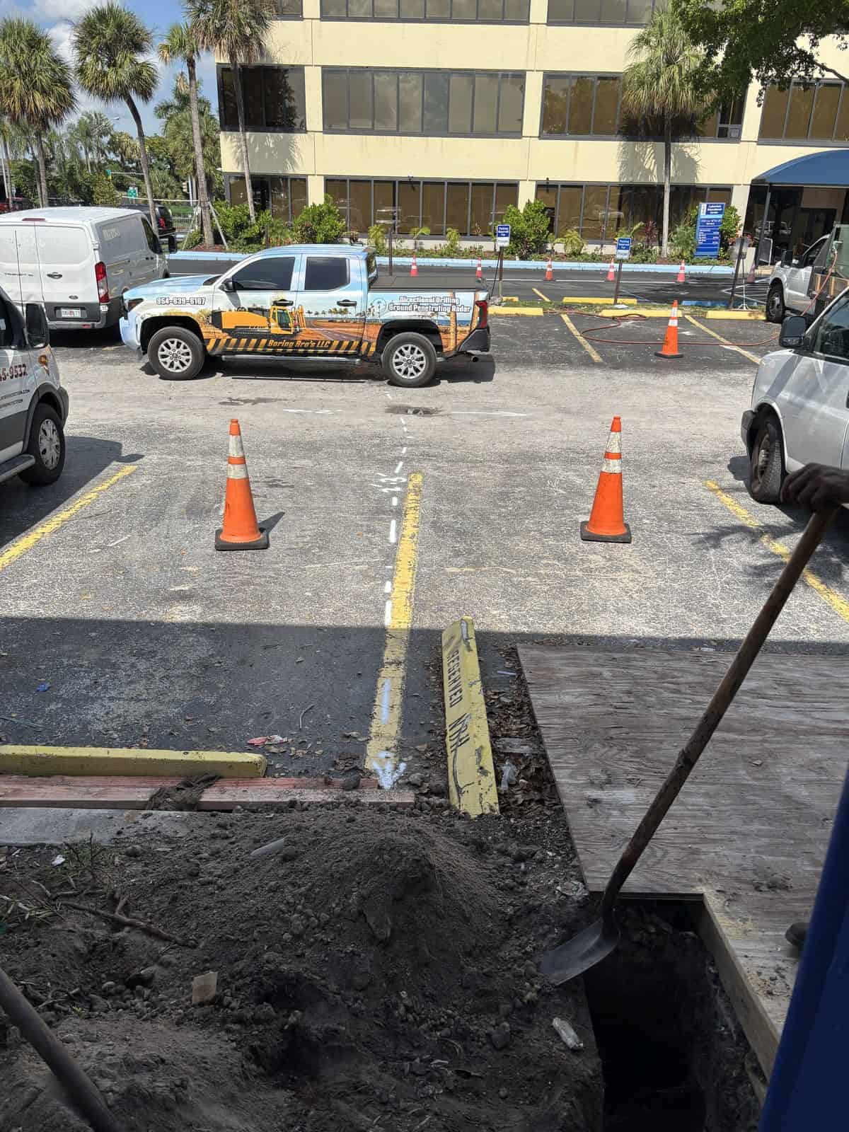 Construction worker digging in parking lot with traffic cones and parked vehicles.