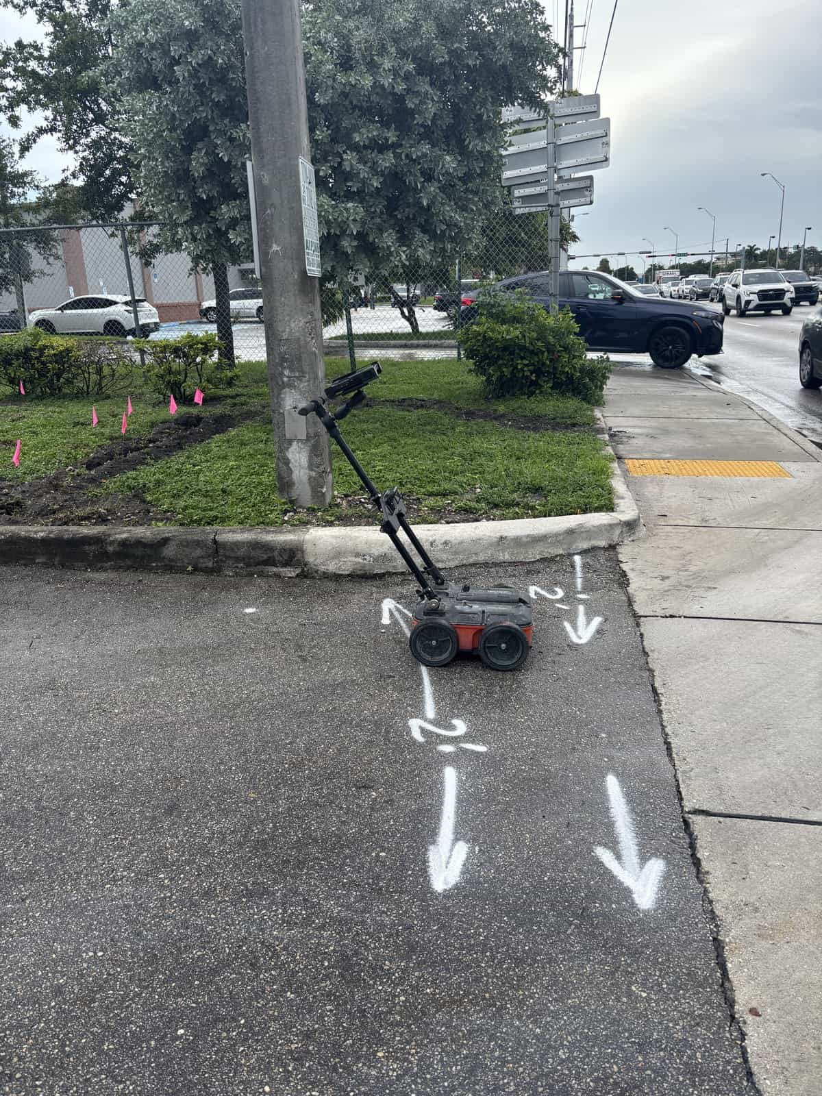 Locating cart on a sidewalk with white arrows and two foot depth marks painted toward a commercial lot entrance in Miami Lakes