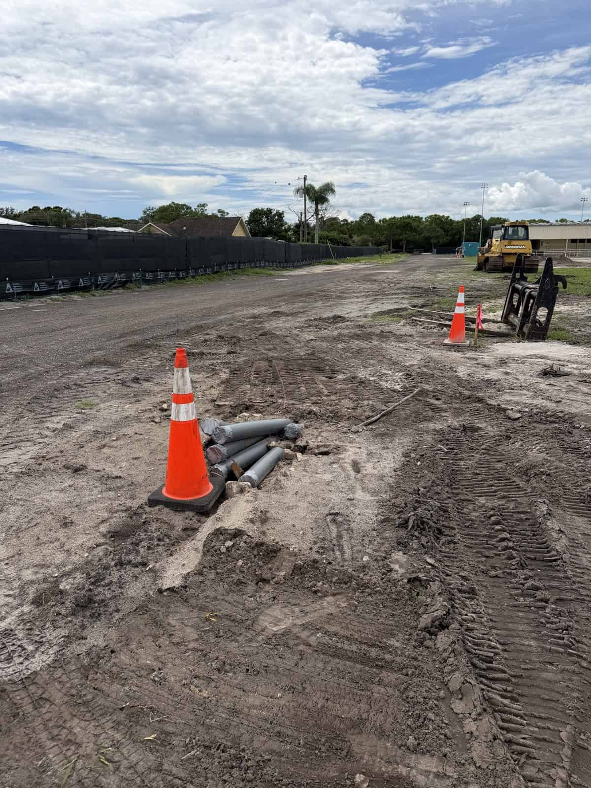 Construction site with orange cones, pipes, and heavy machinery under a cloudy sky.