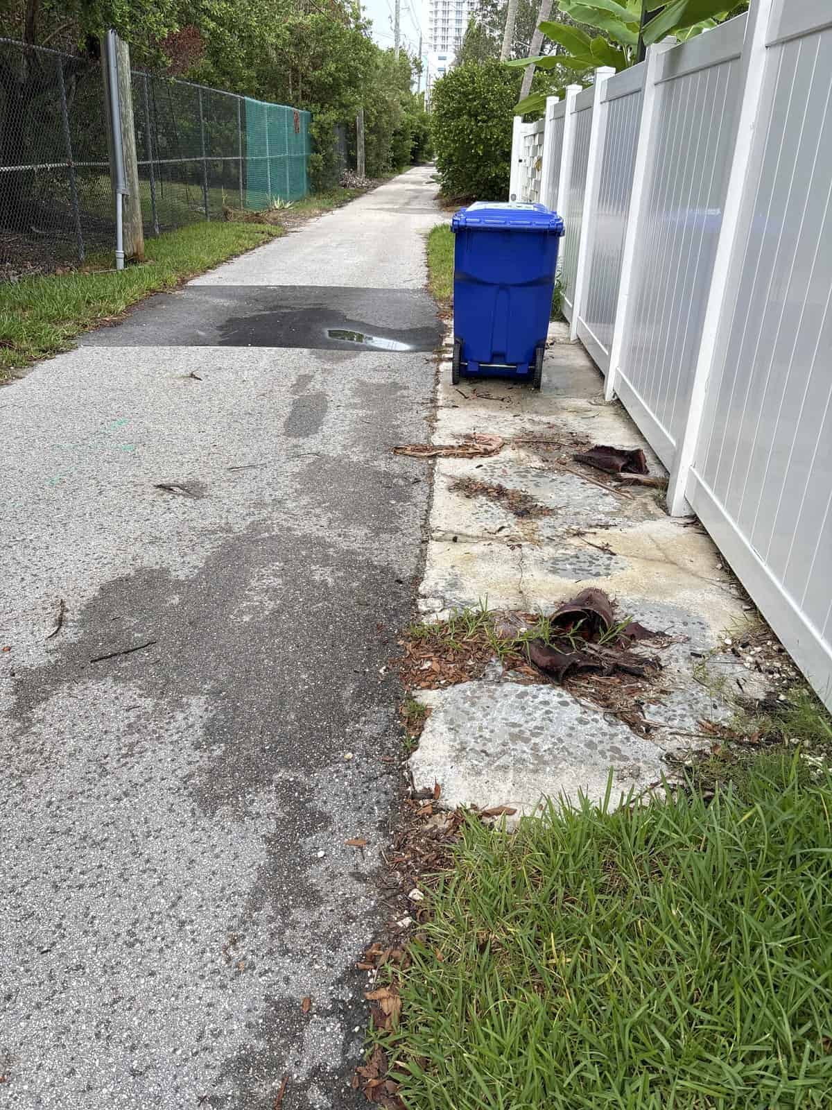 Narrow alleyway with blue trash bin, white fence, and sidewalk damage.