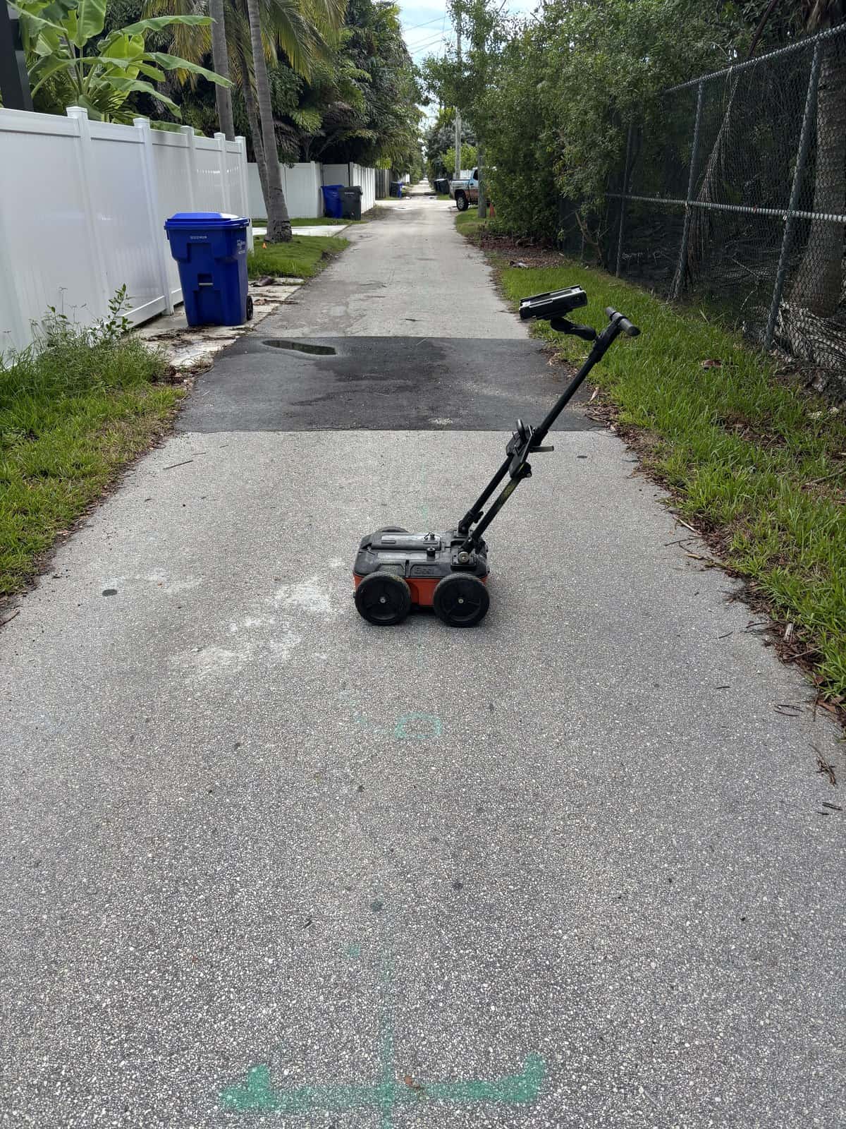 Small robotic surface cleaner on a residential sidewalk with trees and fences.