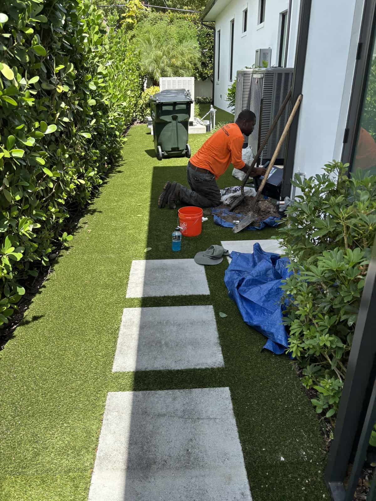 Crew member working by hand in a narrow side yard between air conditioning units and a clipped hedge in Delray Beach