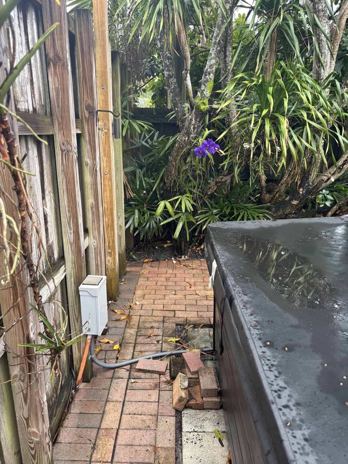 Outdoor hot tub in lush garden with purple flowers and wooden fence.