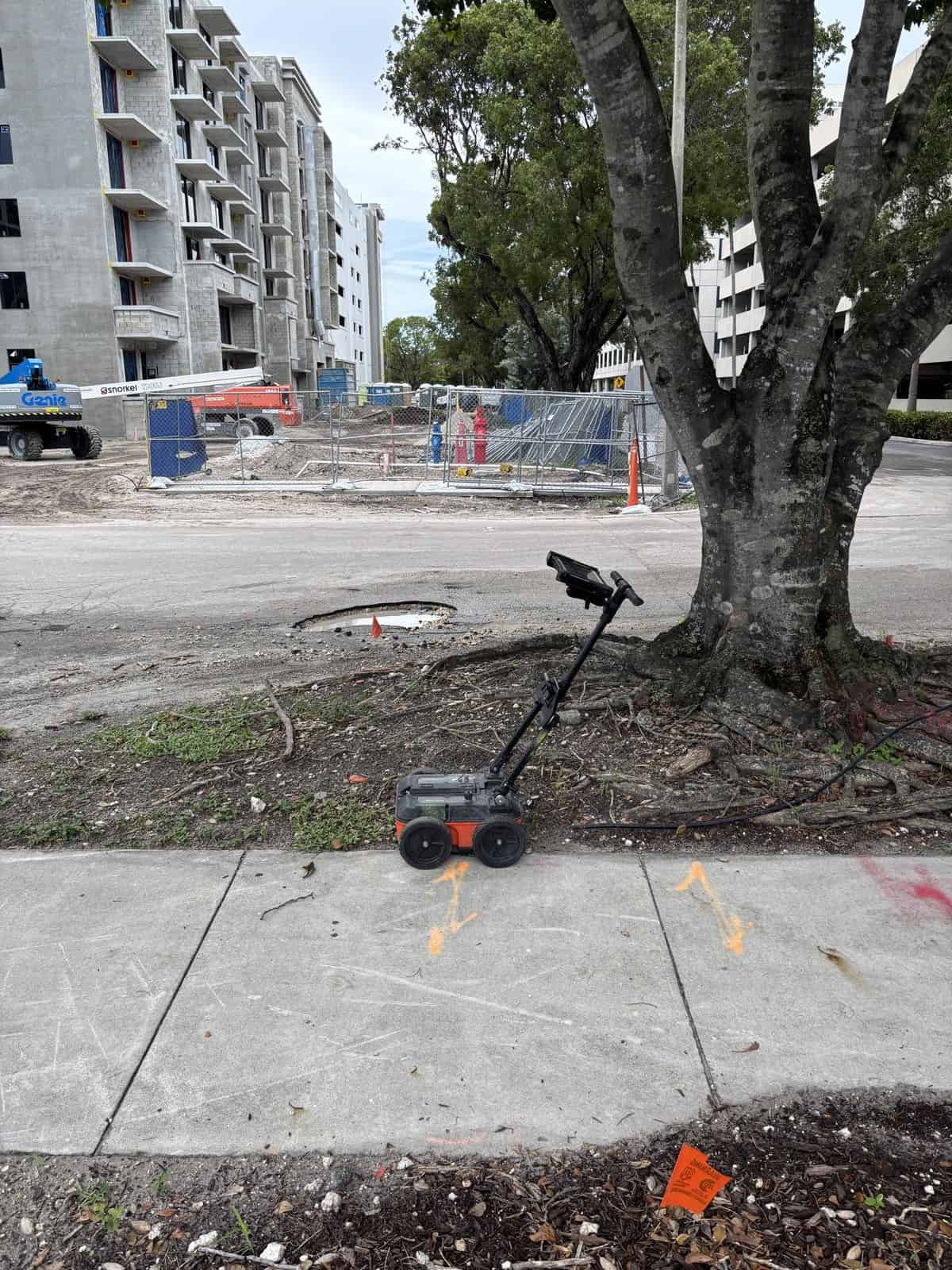 Ground penetrating radar cart on a sidewalk beside a large tree with an active construction site behind it in Fort Lauderdale