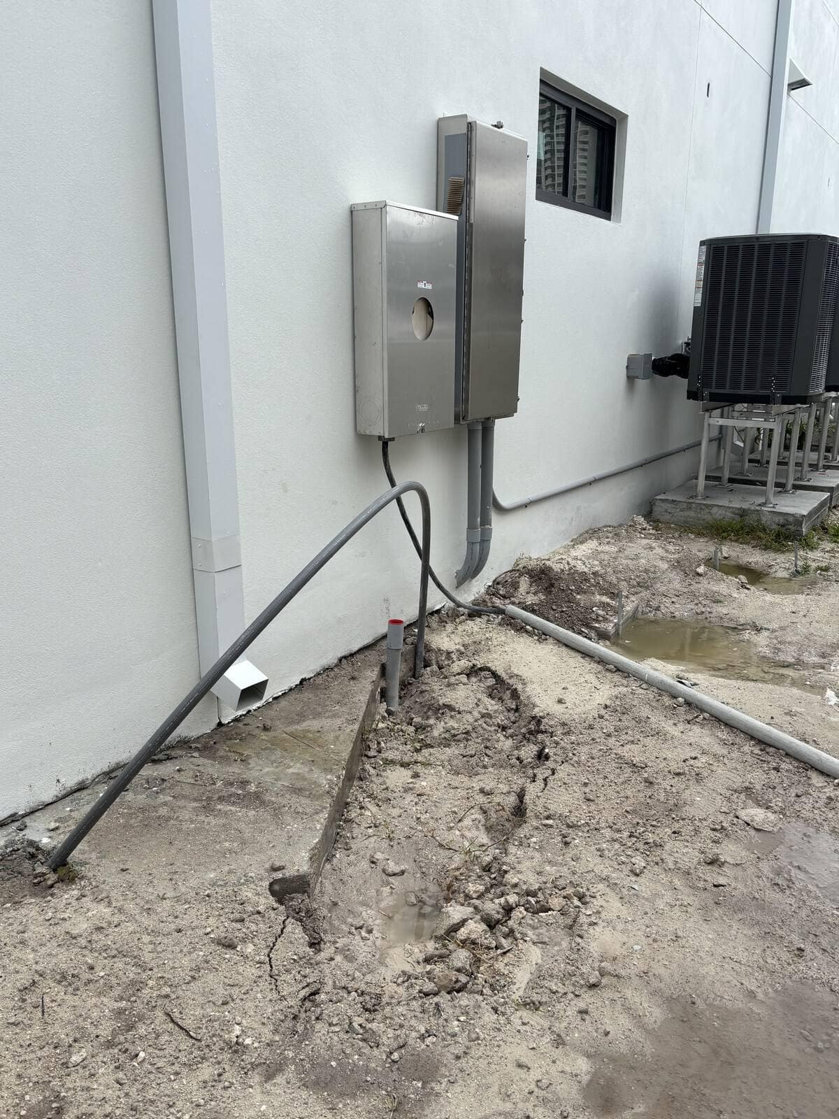 Outdoor electrical boxes and conduit on building exterior with dirt ground.
