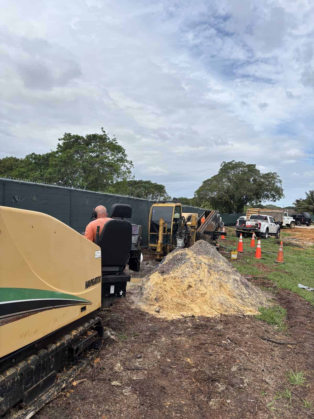 Construction workers and machinery at a site with dirt and orange cones.