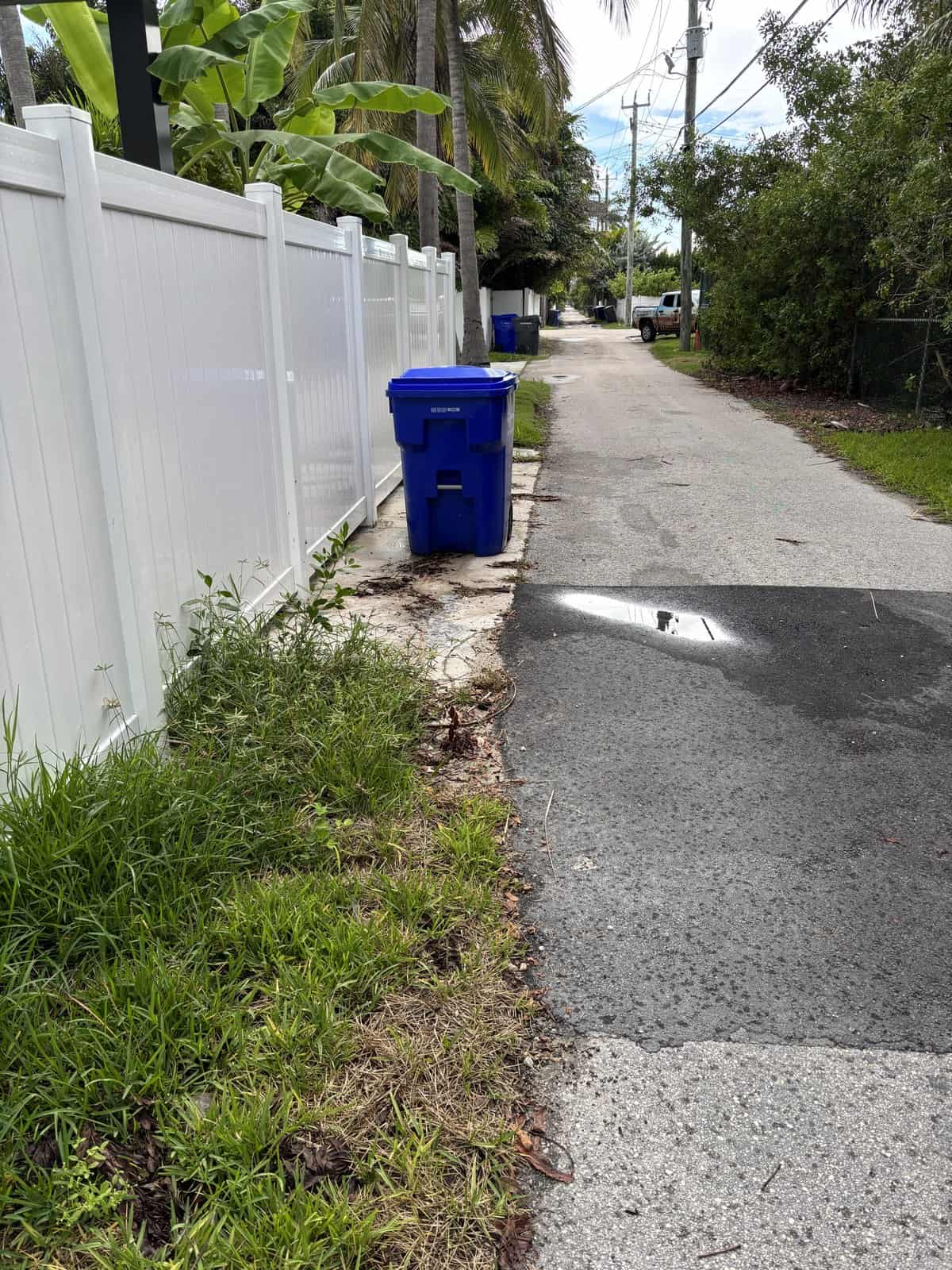 Residential alleyway with white fence, blue trash bin, and greenery.