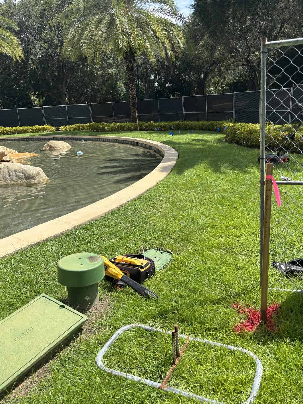 Open green valve box and utility lid on a lawn beside a water feature with locating equipment and a marker flag in Naples